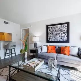 Inviting living room featuring a plush gray sofa with vibrant orange accent pillows, modern artwork, and wood-style flooring. Includes an open breakfast bar with stylish seating and a glass-top coffee table for a bright, contemporary look.
