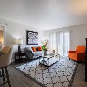 Bright and welcoming living room featuring a gray sofa with orange accent pillows, a coordinating orange armchair, and a glass-top coffee table. Natural light fills the space through large sliding glass doors, complemented by modern décor and wood-style flooring.