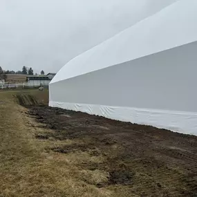 Exterior side view of a long Pilot Buildings white arched fabric structure, positioned parallel to a white fence on muddy ground.