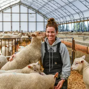 A person smiling while standing among a large flock of sheep inside a well-lit Pilot Buildings arched fabric barn.