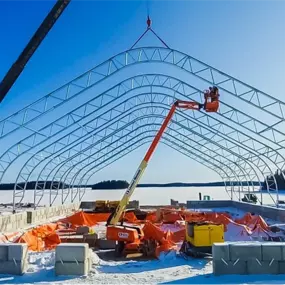 Erection of a Pilot Buildings arched steel frame structure on a concrete block foundation during winter, utilizing a crane and boom lift.