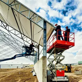 Construction workers on aerial lifts installing the edge section of a white fabric roof cover onto the steel frame of a Pilot Buildings structure built on a concrete wall.