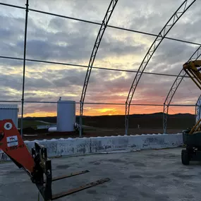 This image showcases the interior of a custom metal building under construction by Pilot Buildings, set against a vibrant sunset. The exposed steel frame and trusses highlight the durable construction of the structure, while the open side reveals a picturesque view of rolling hills. Construction equipment, including a forklift and a boom lift, are visible, indicating an active worksite. The image captures the scale and potential of our custom metal buildings, suitable for various commercial and 