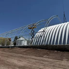 This image captures a large-scale metal arch building, also known as a Quonset hut, during an active construction phase. The photo showcases both the exposed steel truss framework and the section already clad in durable, corrugated metal sheeting. A scissor lift is positioned at the peak of the frame, illustrating the building's impressive height and the ongoing installation process. This provides an excellent view of the robust engineering and quality materials that form our reliable steel buil