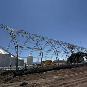 This panoramic image captures a large-scale steel arch building by Pilot Buildings in the midst of construction. The shot effectively displays the building process, with the exposed galvanized steel truss framework in the foreground seamlessly transitioning to the finished, corrugated metal-clad section in the background. Positioned next to a grain silo, the structure's immense length and clear-span design highlight its suitability for major agricultural or industrial applications.