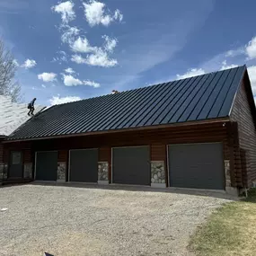 This image showcases a new standing seam metal roof being installed on a beautiful, rustic-style residential garage. The dark metal roof complements the building's log siding and stone accents, demonstrating a blend of durability and high-end aesthetics. With workers actively fitting the panels, this photo highlights our expert installation services for residential projects. This custom four-car garage is a prime example of how our metal building expertise extends to custom homes and workshops.