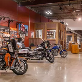 The image shows the interior of Mike's Famous Harley-Davidson, featuring polished floors, industrial-style ceilings, and a lineup of gleaming motorcycles, including orange, silver, and blue models. The background displays branded parts and accessories, with signage and an American flag adding to the Harley-Davidson aesthetic. A rustic wooden counter enhances the space's vintage charm.