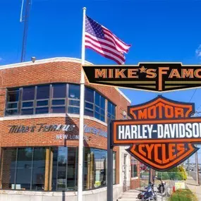 This image features the exterior of Mike's Famous Harley-Davidson, housed in a historic brick building with large, curved glass windows. The American flag waves prominently above the dealership, emphasizing its patriotic and iconic branding. In the foreground, the Mike's Famous logo is displayed above the classic Harley-Davidson 