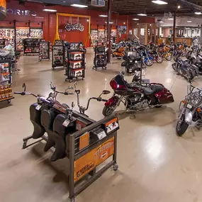 This image showcases the expansive interior of Mike's Famous Harley-Davidson, featuring a large showroom with polished concrete floors. The space is filled with a wide variety of Harley-Davidson motorcycles, including vibrant models in orange, red, and silver, all neatly arranged for display.

The background highlights shelves stocked with branded merchandise and accessories, with clear organization and signage. In the center, display carts showcase products like handlebars, seats, and other par