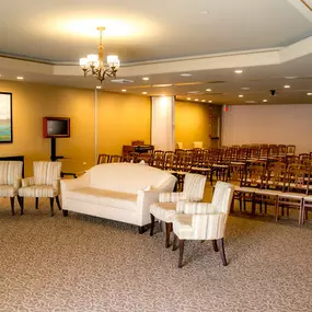 This image captures the interior of the chapel at Brady-Gill Funeral Home & Cremation Services. Rows of elegant wooden chairs are neatly arranged to accommodate guests during a service. At the front, a cozy seating area with a cream sofa and matching armchairs provides a welcoming space for close family. Warm tones, soft lighting, and tasteful decor create a serene and comforting ambiance.