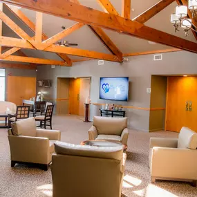 This image showcases the inviting lobby of Brady-Gill Funeral Home & Cremation Services. The space features comfortable armchairs and a small round table arranged for guests to gather. Exposed wooden beams add rustic charm, while chandeliers and warm lighting create an elegant ambiance. A flat-screen TV displaying the Brady-Gill logo adds a modern touch to the classic design.