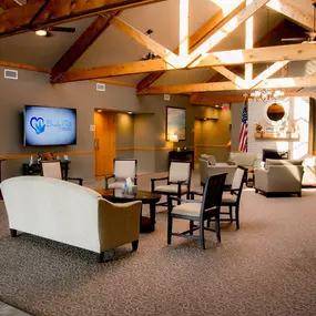 This image showcases the spacious and welcoming lobby of Brady-Gill Funeral Home & Cremation Services. The area features various seating arrangements with chairs and sofas, creating a comfortable space for families and visitors. Exposed wooden beams, a large flat-screen TV displaying the Brady-Gill logo, and elegant lighting enhance the lobby's warm and professional atmosphere. A brick fireplace and thoughtful decor add to its charm.