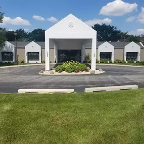 The image displays the exterior of Brady-Gill Funeral Home & Cremation Services in Tinley Park, IL. The white building features a prominent covered entrance with clean, modern lines, surrounded by neatly landscaped greenery and a circular driveway for easy access. The bright blue sky and lush trees in the background add to the serene and welcoming atmosphere.