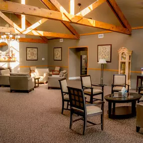 This image captures a cozy sitting area within the lobby of Brady-Gill Funeral Home & Cremation Services. The space features a mix of comfortable chairs and sofas arranged in small conversational groupings. Exposed wooden beams, soft lighting, and elegant decor create a warm and inviting atmosphere. A brick fireplace and an American flag add a sense of tradition and respect, while artwork and a grandfather clock enhance the room’s charm.