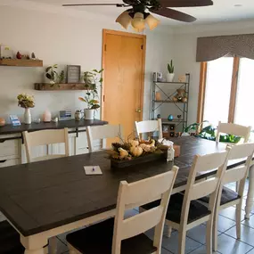 This image showcases a private meeting room at Brady-Gill Funeral Home & Cremation Services. The room features a rustic wooden table surrounded by white chairs, creating an inviting space for consultations or family discussions. Decorated with plants, shelves displaying keepsakes, and a calming neutral color palette, the room is filled with natural light from large windows that enhance its warmth and tranquility.