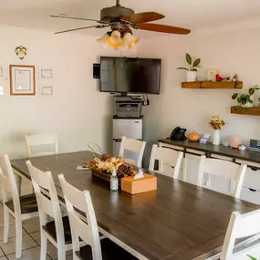 This image highlights another view of the private meeting room at Brady-Gill Funeral Home & Cremation Services. The space features a large rustic wooden table with white chairs, ideal for consultations or family discussions. A wall-mounted television and document display area add functionality, while the shelves with plants and decor create a warm and welcoming atmosphere. Certificates on the wall reflect professionalism and dedication to service.