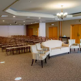 This image features the interior of the chapel at Brady-Gill Funeral Home & Cremation Services. Rows of wooden chairs are neatly arranged for guests, providing ample seating for a memorial service. At the front of the room, a comfortable seating area with a cream-colored sofa and matching armchairs is arranged for family members. The soft lighting, neutral tones, and tasteful design create a serene and respectful atmosphere.