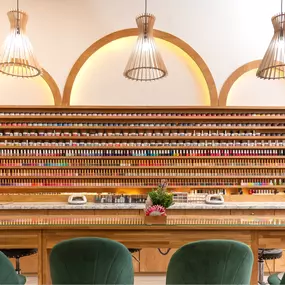 This image perfectly captures the luxurious and meticulously designed interior of Eighty-8 Nail Studio. The focus is on an impressive, wall-to-wall display of nail polish bottles, beautifully arranged by color and style, showcasing the studio's extensive selection of premium nail products. The shelving, crafted from warm wood with elegant arch accents, highlights the studio’s modern yet inviting aesthetic.  

A marble countertop beneath the display adds a touch of sophistication, while neatly ar