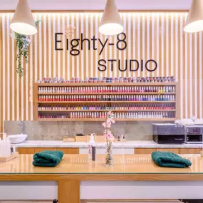 This image showcases the refined and stylish workspace of Eighty-8 Studio, emphasizing its commitment to delivering a high-quality and luxurious beauty experience. The focal point is a beautifully arranged nail polish display against a modern wooden slatted wall, combining functionality and aesthetic appeal. The wide range of nail colors on display reflects the studio’s extensive offerings and dedication to customer choice.  

A sleek countertop with a glass surface, adorned with neatly folded g