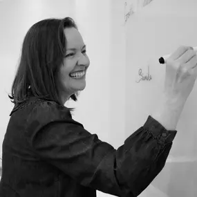 Lisa Powell is laughing as she writes on a whiteboard during a lesson, demonstrating her interactive and approachable teaching style. Her connection with students is evident in her cheerful demeanour.