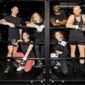 A group of six Jabz Boxing members pose confidently inside the custom boxing ring. Smiling and wearing gloves, they showcase the gym’s welcoming and supportive community. The boxing bags and gym setup in the background emphasize the fitness environment.