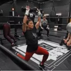 A group fitness class in progress at Jabz Boxing. Participants are engaged in a variety of exercises, including a woman in the foreground performing a lunge with dumbbells overhead. The custom boxing ring with the Jabz logo on the mat serves as the centerpiece, creating an energetic and focused environment.