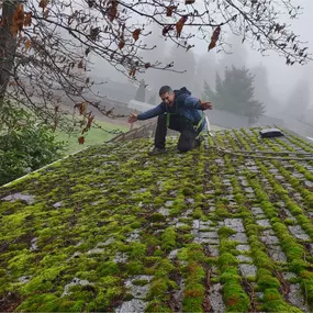 A man kneels on a mossy roof, indicating the need for cleaning and maintenance before restoration.