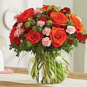 A vibrant bouquet of orange roses, red carnations, pink mini carnations, and lush greenery beautifully arranged in a round glass vase. The floral centerpiece is displayed on a wooden dining table, creating a warm and inviting atmosphere, ideal for fall celebrations, romantic occasions, or as a stunning Thanksgiving decoration. The neutral background enhances the bold colors of the flowers, making them the focal point of the setting