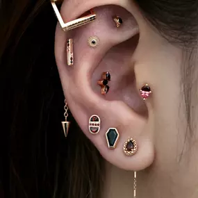 A close-up of an ear with multiple rose gold earrings, featuring geometric shapes, dark gemstones, and chain details.