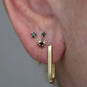 A close-up of an ear with black gemstone earrings, including a spider-shaped piece with a dangling chain and a gold hoop.