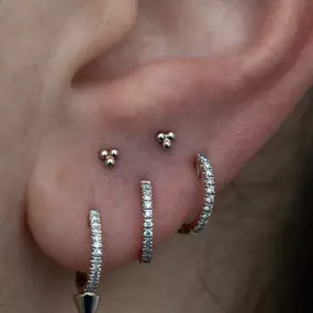 A close-up of an ear with silver jewelry, featuring two small trinity studs and three diamond-encrusted hoops, one with a pointed tip. The piercings are arranged along the lobe.