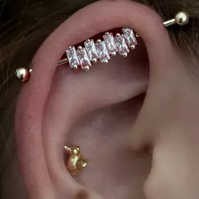 A close-up of an ear with an industrial piercing featuring a barbell adorned with pink baguette-cut gemstones. Additional gold earrings include a small duck and a key design.