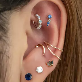 An ear decorated with mixed metal jewelry, including a silver opal cuff, a blue gemstone drop earring, a curved gold spike, and various studs