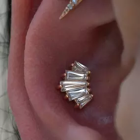 A close-up of an ear with gold jewelry, including a lightning bolt stud, a baguette-cut gemstone cluster in the conch, and a small diamond stud below.