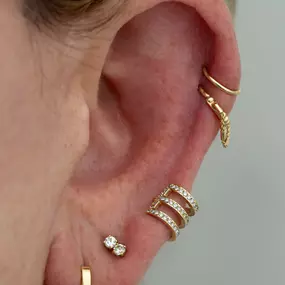 An ear with multiple gold earrings, including a triple-band diamond cuff, a small diamond stud, and decorative helix cuffs.