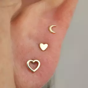 A close-up of an ear with three gold earrings: a crescent moon, a solid heart, and an open heart stud arranged vertically.