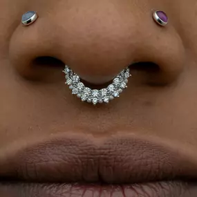 A close-up of a nose with a white gold septum ring encrusted with diamonds, along with two symmetrical nostril piercings featuring bezel-set gemstones in blue and purple.