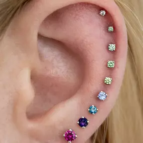 An ear with a gradient of colorful gemstones, arranged from small green stones on the helix to larger pink, purple, and blue studs on the lobe.