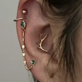 An ear with multiple gold and green gemstone earrings, including a chain earring, a cuff, a curved antler-shaped piece, and a helix piercing with a gold bead.