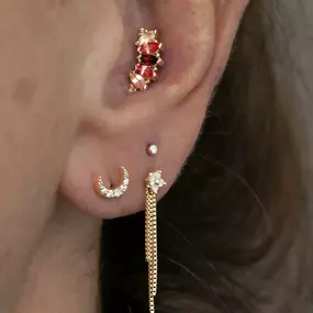 A close-up of an ear adorned with gold jewelry, including a red and peach gemstone cluster on the helix, a tiny stud, a crescent moon earring, and a long chain earring with a floral accent on the lobe.