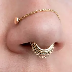 A close-up of a nose with gold jewelry, featuring a septum ring with layered chains and black gemstones, paired with a nostril piercing connected to a braided gold chain.