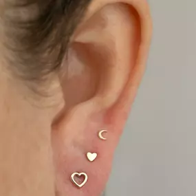 Side view of an ear with gold heart-themed earrings, including a crescent moon, solid heart, and open heart studs.