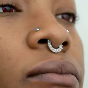 A side profile view of a person with a silver diamond-encrusted septum ring and two nostril piercings with bezel-set gemstones. The eyes are slightly visible in the frame.