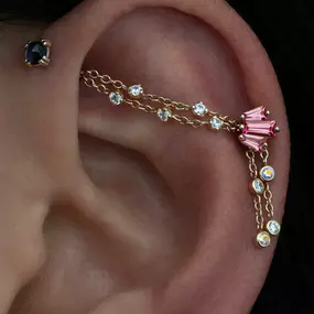 An ear with a delicate gold chain earring connecting two piercings, featuring a pink baguette cluster and sparkling bezel-set diamonds.