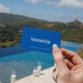 The photo shows a hand holding an Instadrip business card with the words “Concierge IV Infusion Therapy,” in front of a bright blue pool. The background includes green hills and cylindrical white candles by the water’s edge.