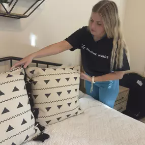 This image showcases a Breathe Maids cleaning professional meticulously arranging decorative pillows on a neatly made bed. She is dressed in a Breathe Maids branded black t-shirt, reinforcing the company's professional image. The scene exudes a sense of cleanliness, organization, and attention to detail, which aligns with the brand’s commitment to high-quality home cleaning services.

The modern, cozy interior setting—with a stylish shelf, neutral tones, and well-curated décor—further emphasizes