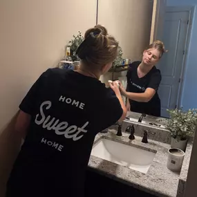 This image beautifully showcases a Breathe Maids of Orlando cleaning professional meticulously wiping down a bathroom mirror and sink area. She is wearing a branded black t-shirt with the words 