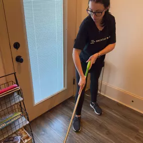 This image captures a Breathe Maids cleaning professional hard at work, ensuring a spotless and well-maintained home. She is wearing a Breathe Maids branded black t-shirt, reinforcing the company’s professional and polished look. Focused and detail-oriented, she is mopping the hardwood floor near a bright entryway, demonstrating the company’s commitment to thorough, high-quality cleaning services.

The cozy home setting, with neatly organized storage shelves and a welcoming atmosphere, highlight