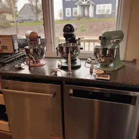 Colorful KitchenAid stand mixers on display in a kitchen window setup, showcasing multiple finishes and attachments.