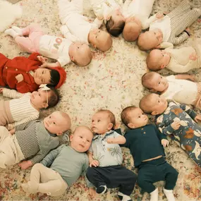 Fourteen babies lay in a circle smiling and cooing on a floral carpet